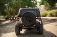 Back view of a white 2007-2018 Jeep JK 4-Door with DV8 Fastback Hard Top with a spare tire on a dirt road surrounded by trees.