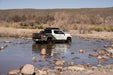 Tacoma crossing a shallow river with rocky bed and desert landscape in the background