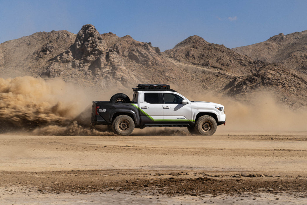 White Tacoma with DV8 roof rack driving on a dirt road in a desert landscape with mountains in the background.